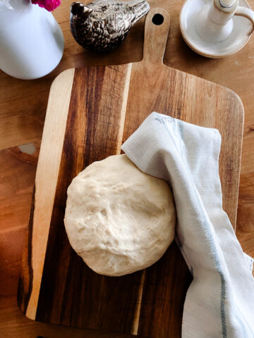 homemade pizza dough on a wood cutting board next to a tea towel